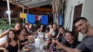 Een groep mensen zit aan tafel met verschillende gerechten en drankjes, onder een overdekt terras met planten en kleding op de achtergrond.