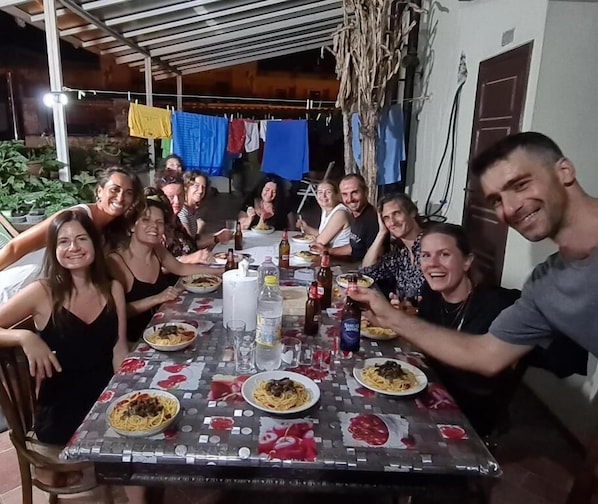 Un grupo de personas sentadas alrededor de una mesa con diversos platos y bebidas, bajo un espacio al aire libre cubierto con plantas y ropa colgada al fondo.