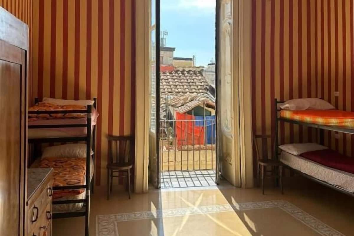 Une pièce aux murs rayés, avec un lit superposé, un bureau et une vue sur l’extérieur par une porte ouverte.