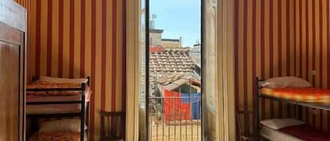 Une pièce aux murs rayés, avec un lit superposé, un bureau et une vue sur l’extérieur par une porte ouverte.