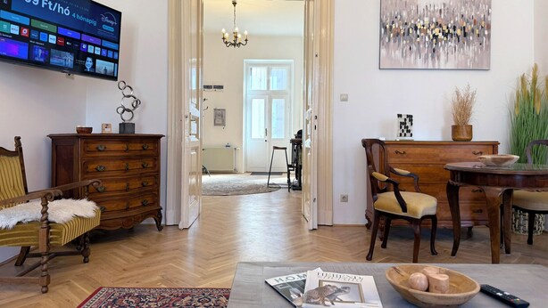 Una sala de estar con televisor mostrando un servicio de streaming, un aparador de madera, un sillón con una manta de piel, una mesa de comedor con sillas y un cuadro en la pared.