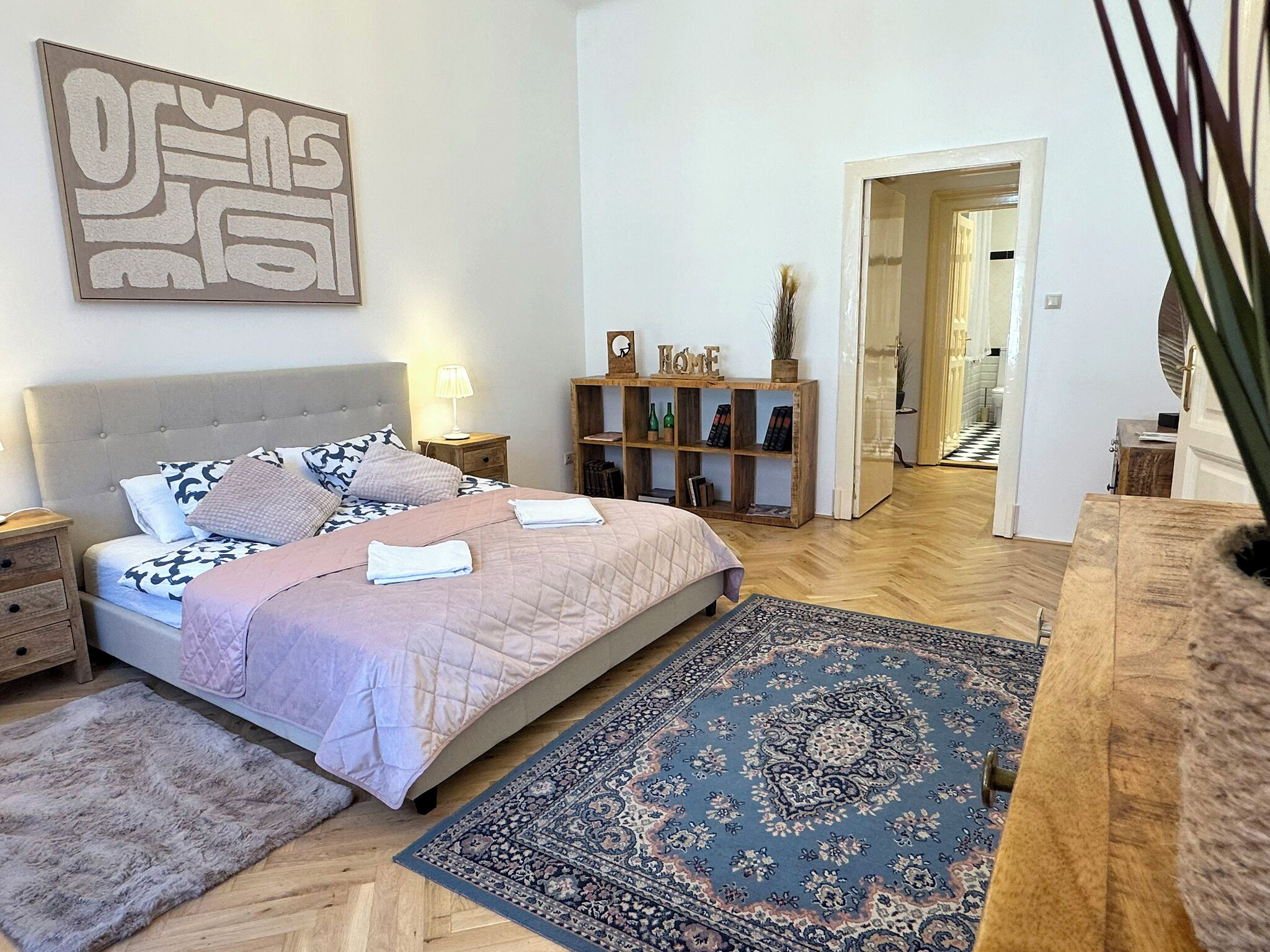 Een slaapkamer met een groot bed, een nachtkastje, een houten bijzettafeltje, een wandplank met decoratieve objecten en een kamerbreed tapijt.