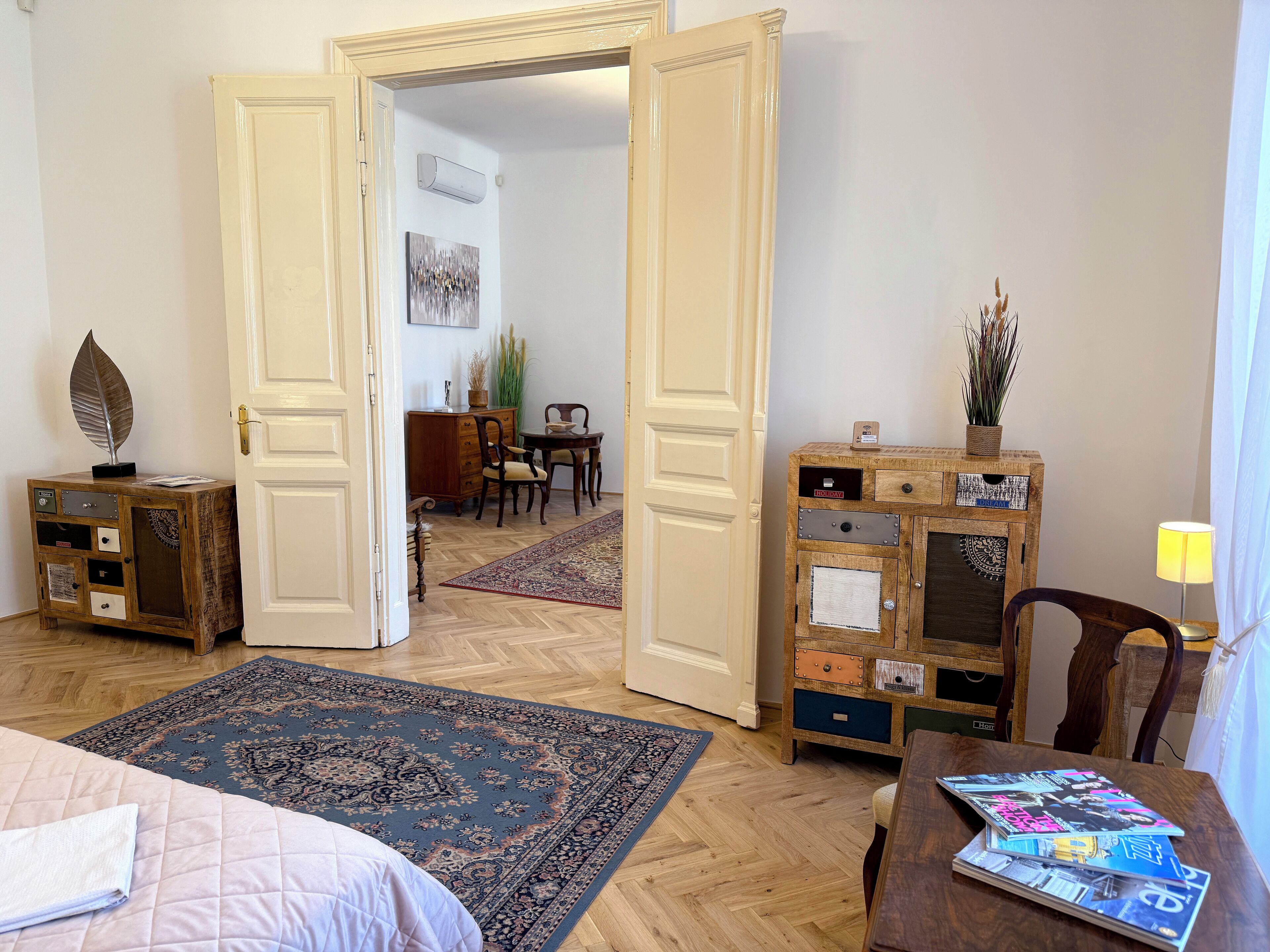 A hotel room with a bed, a wooden chest of drawers, a small table with magazines, a rug, and a door leading to another room.