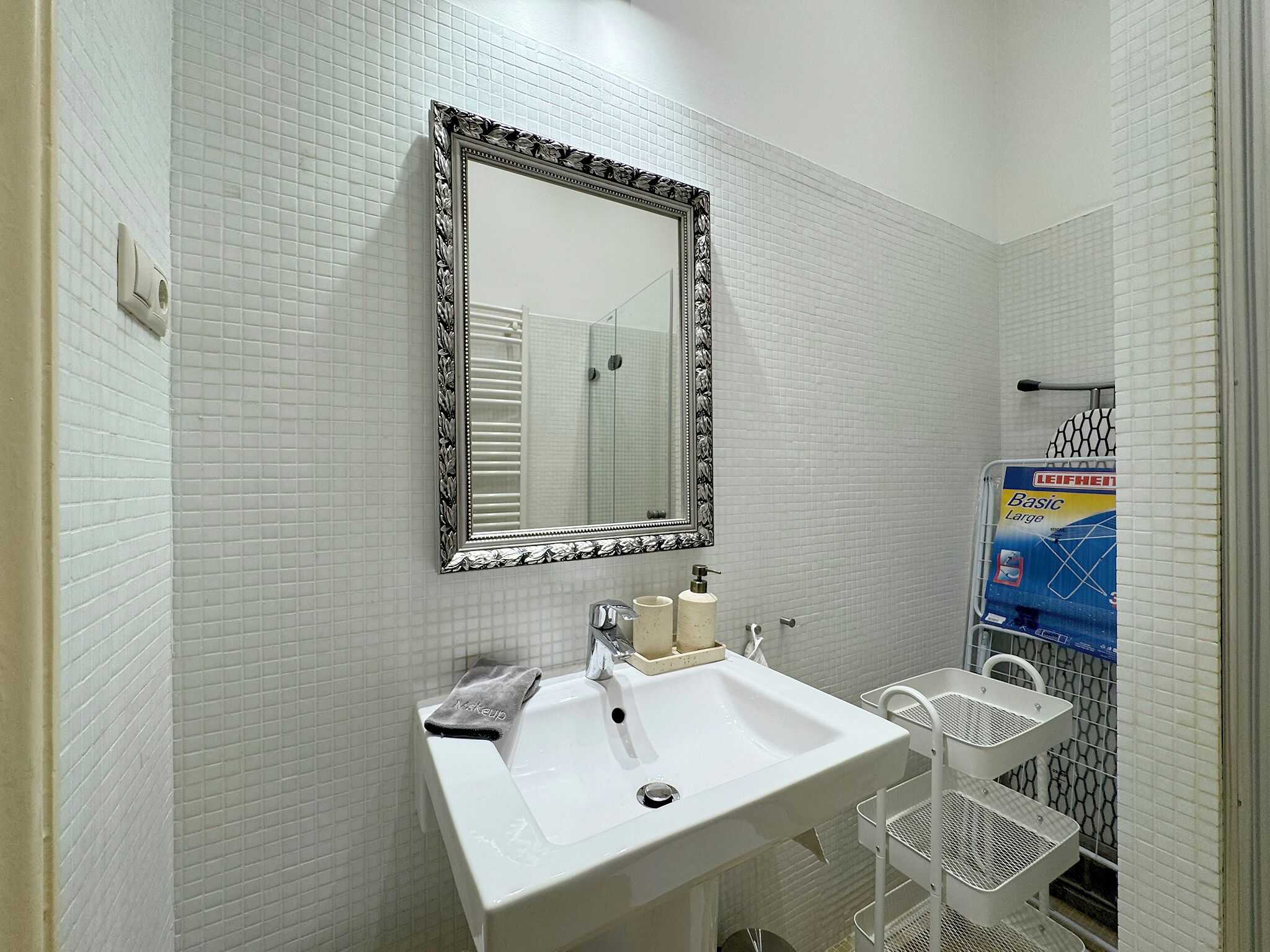 A bathroom with a white sink, a mirror with a decorative frame, a towel, and a Leifheit shower caddy.