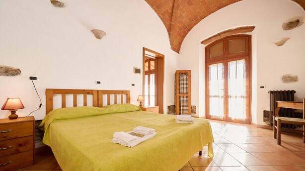 A bedroom with a bed, a nightstand, a lamp, a wooden chair, and a window with curtains.