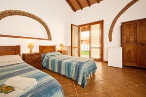 Une chambre avec deux lits, une armoire en bois, un petit réfrigérateur et une porte donnant sur un balcon.
