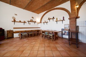 Une pièce avec une longue table en bois, plusieurs chaises, une cheminée en briques, des étagères ornées d’objets décoratifs et un sol carrelé.