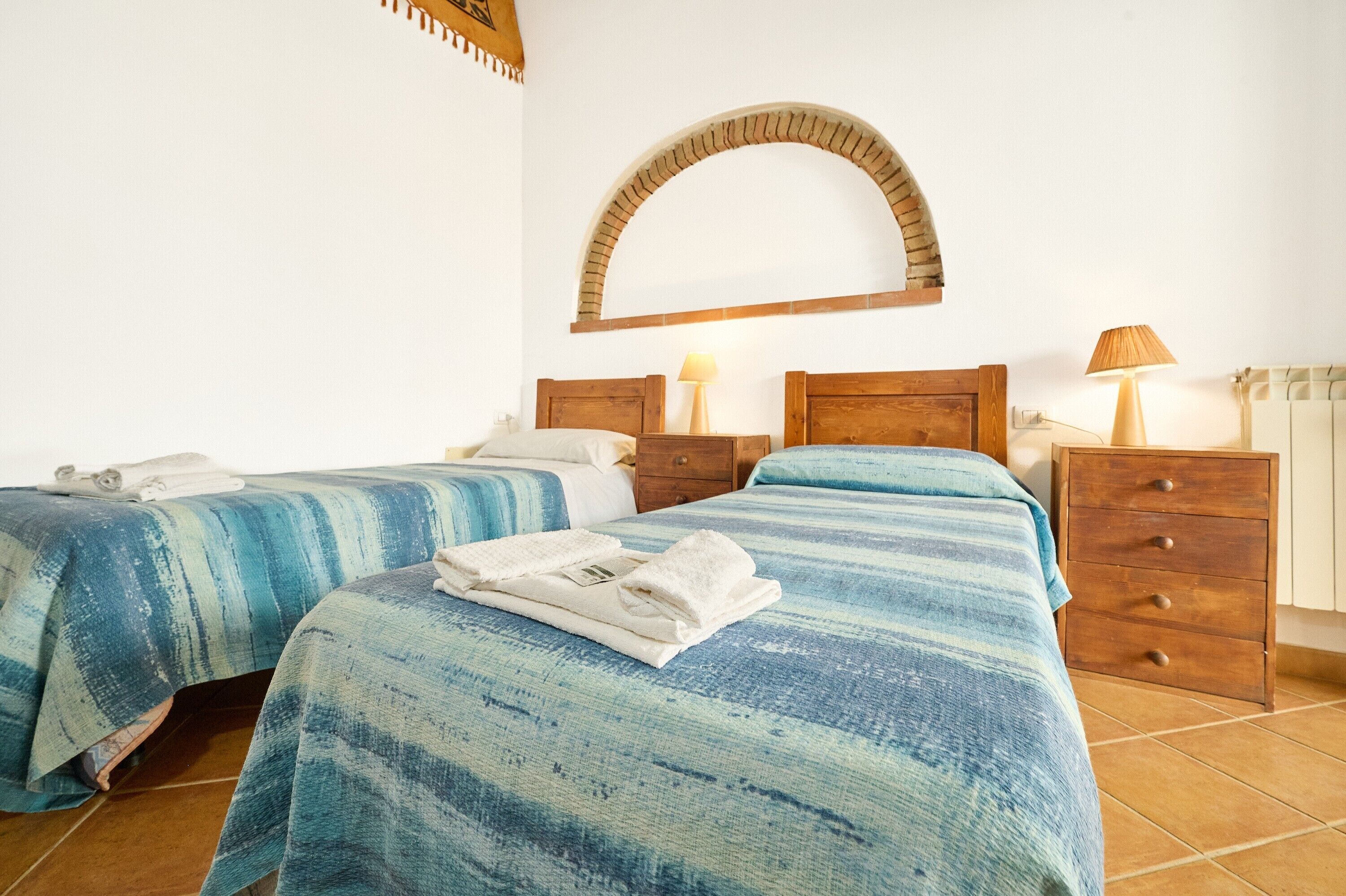 Habitación de hotel con dos camas, cabeceros de madera, suelo de baldosas y un espejo arqueado montado en la pared.