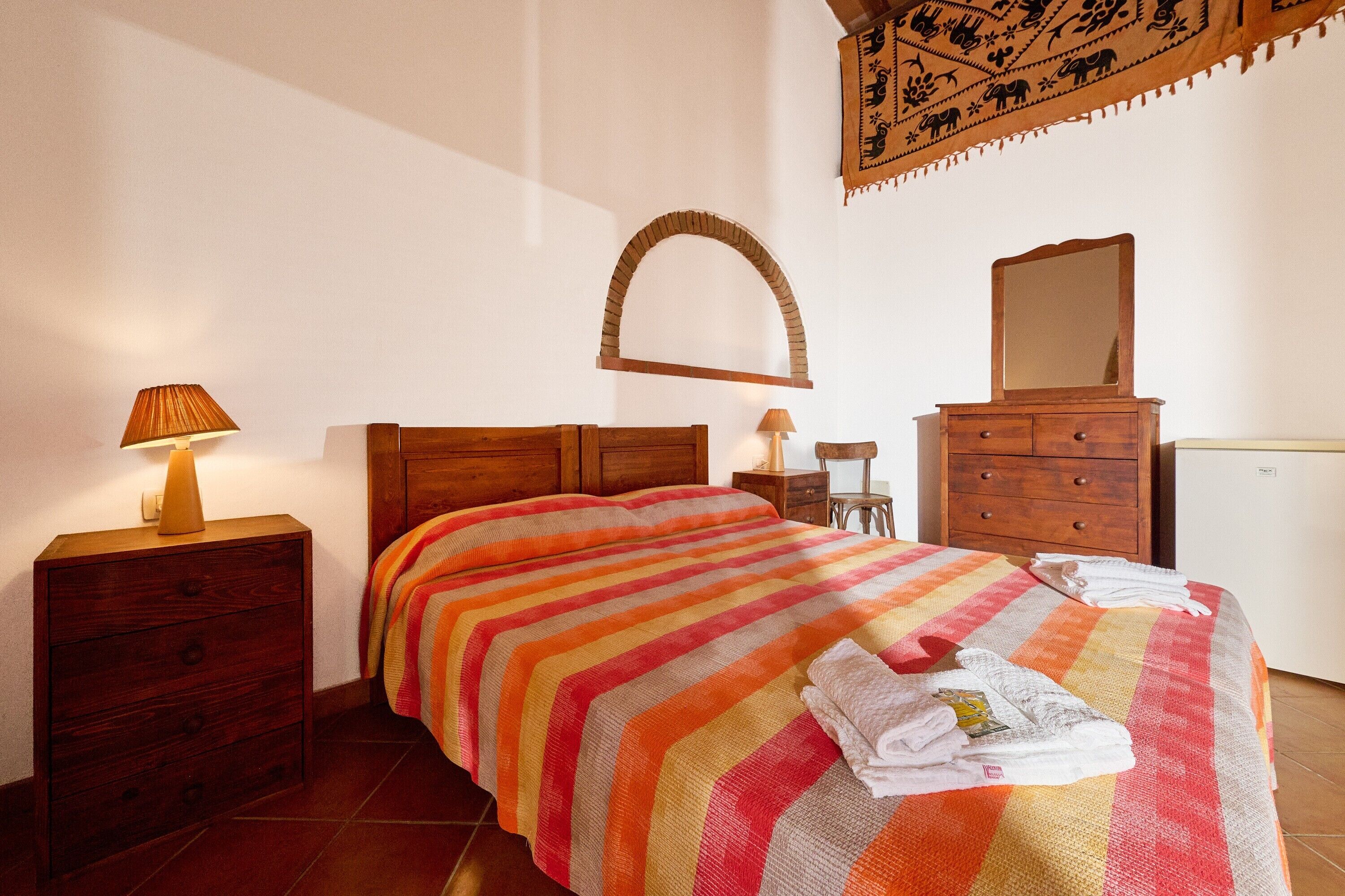 Een slaapkamer met een bed, een houten dressoir, nachtkastje, lamp en een spiegel.