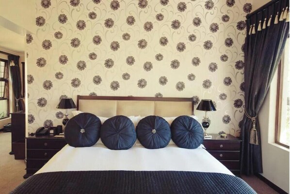 Una habitación de hotel con una cama grande, ropa de cama azul oscuro y un estampado de papel tapiz floral.