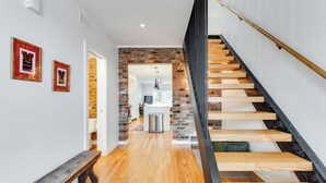 Un escalier avec des marches en bois et une rampe noire, menant à une pièce dont un mur est en briques et la porte est blanche.