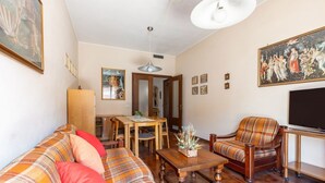 En stue med sofa, lænestol, sofabord i træ og et fjernsyn. Der er et spisebord med stole og et billede på væggen.