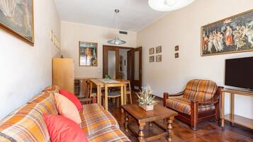 Una sala de estar con sofá, sillón, mesa de centro de madera y televisor. Hay una mesa de comedor con sillas y un cuadro en la pared.