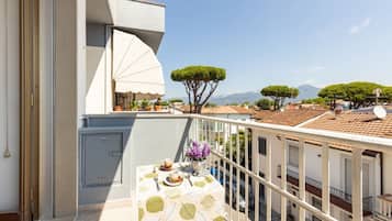 Un balcón con una mesa dispuesta para dos personas, una sombrilla blanca y vistas a árboles y edificios.