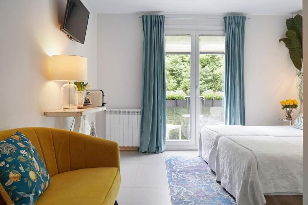A hotel room with a bed, a sofa, a TV, a window with curtains, and a view of greenery outside.