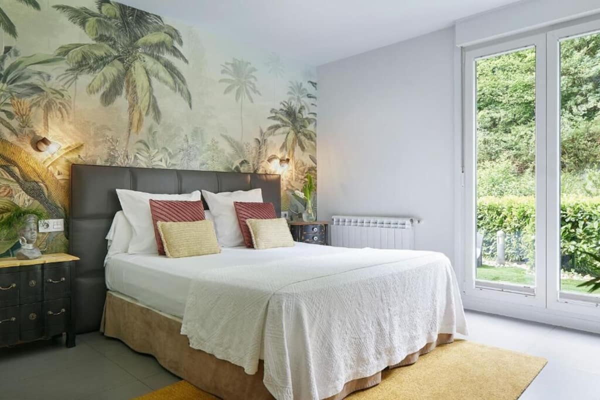 A bedroom with a large bed, a nightstand, a dresser, and a window with a view of greenery.