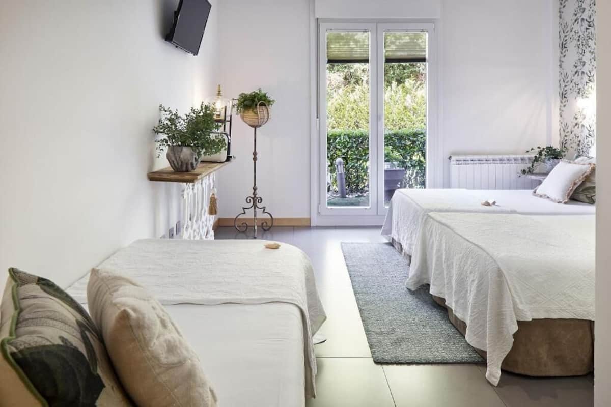A bedroom with two beds, a small table with a plant, a floor lamp, and a view of the outdoors through a sliding door.
