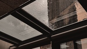 Une lucarne avec des gouttes de pluie sur la vitre, un cadre en métal et une vue sur un bâtiment à l’extérieur.