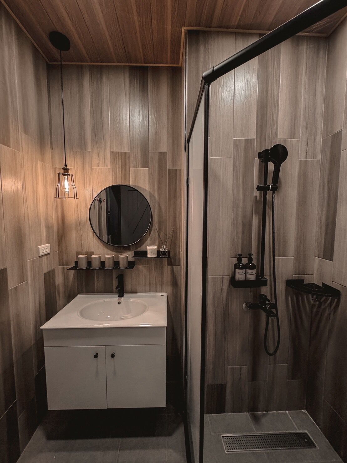 A modern bathroom with a wooden panel wall, a round mirror, a white sink, and a shower area.