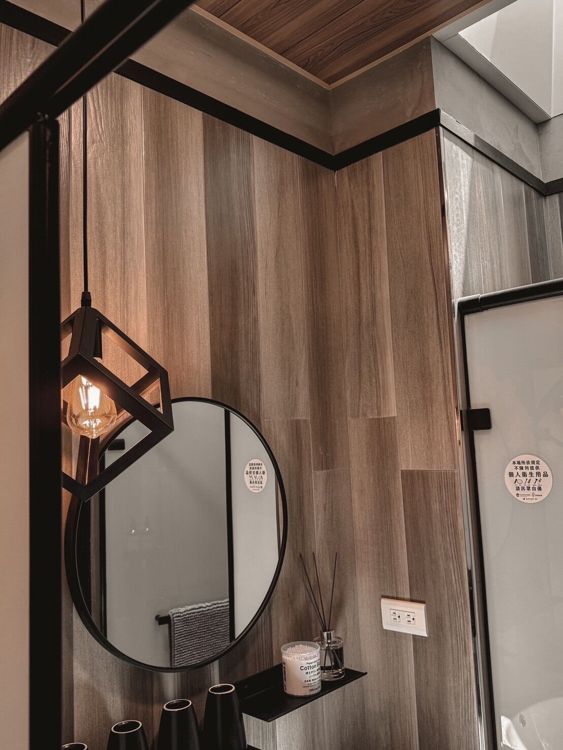 A bathroom with a round mirror, wooden panel walls, a hanging light fixture, and a shelf with toiletries.