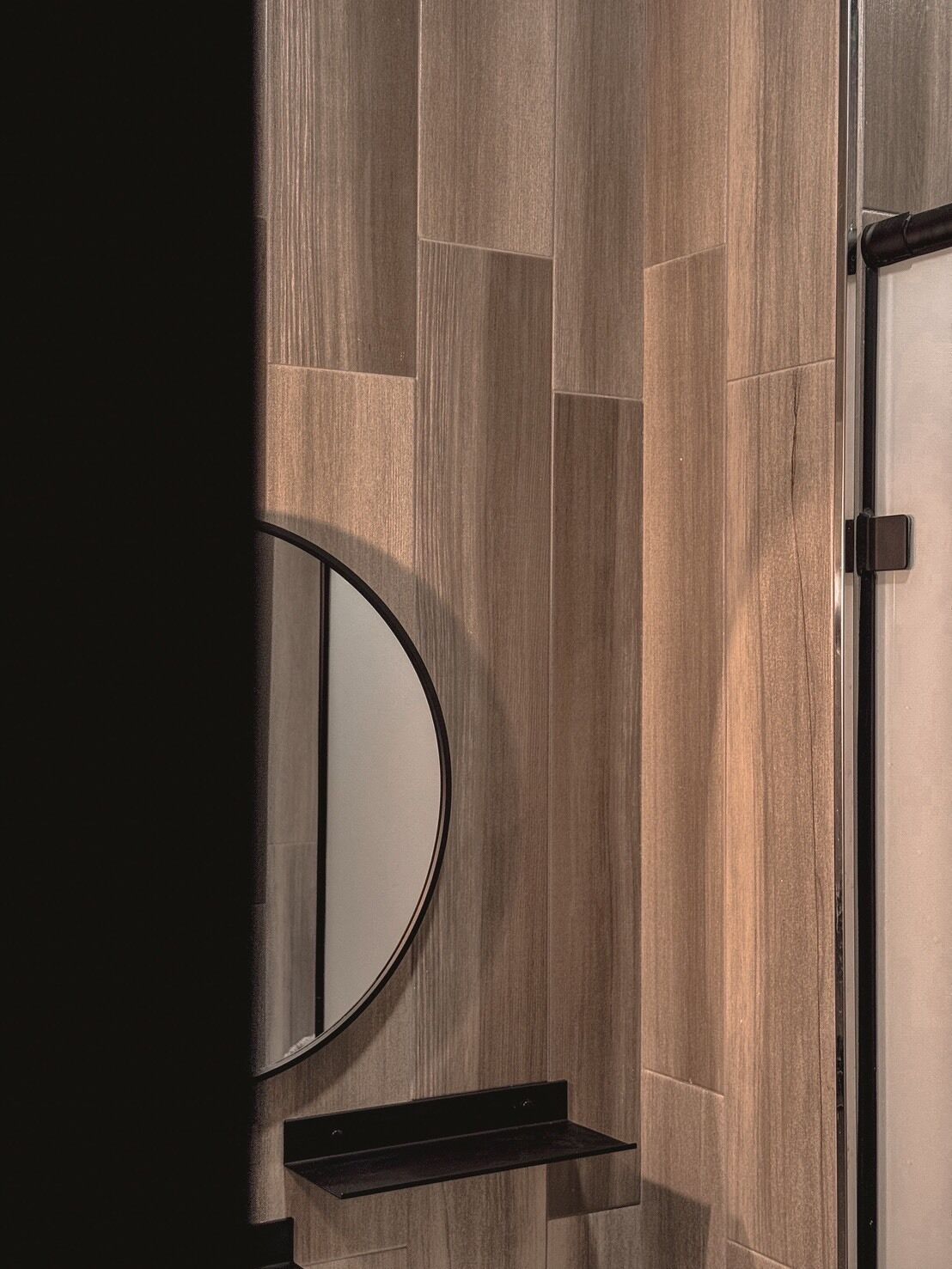 Un baño con pared de madera, un espejo redondo y un estante negro.