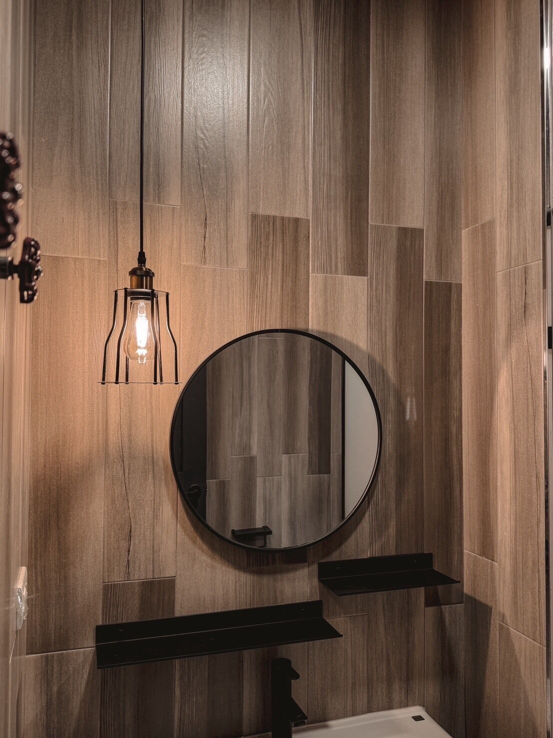 A bathroom with a wooden wall, a round mirror, and a hanging light fixture.