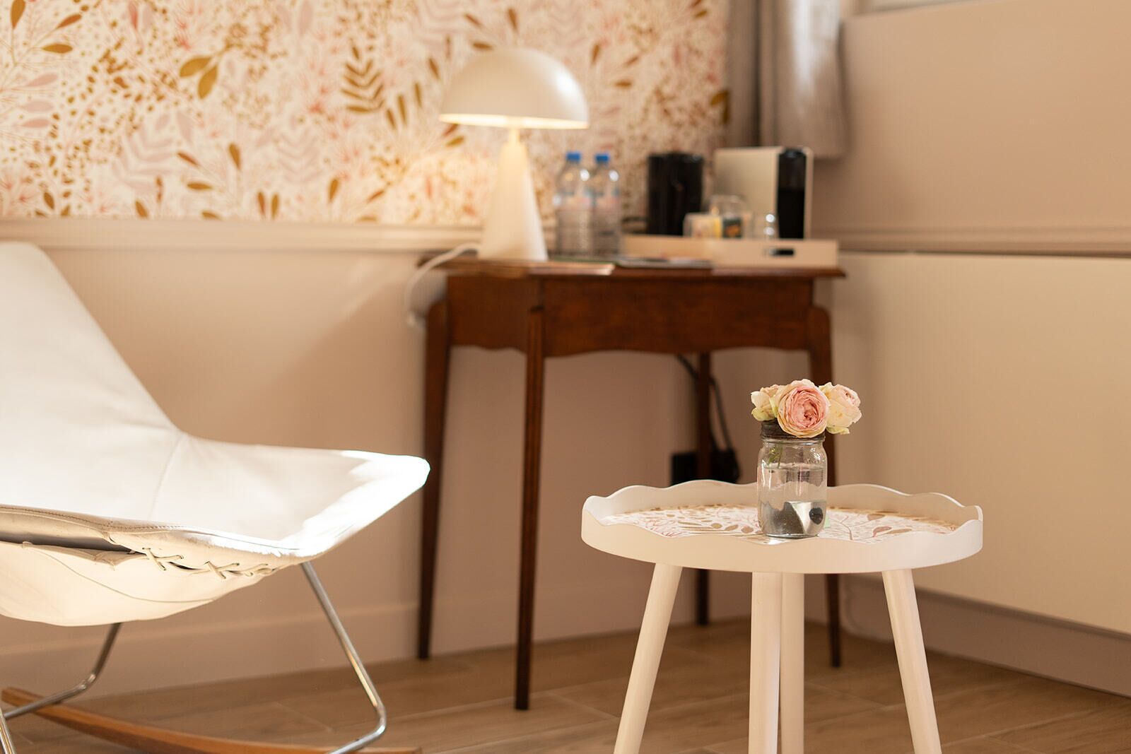 A cozy room with a white chair, a small round table with a vase of flowers, a lamp, and a wooden desk with a coffee maker.
