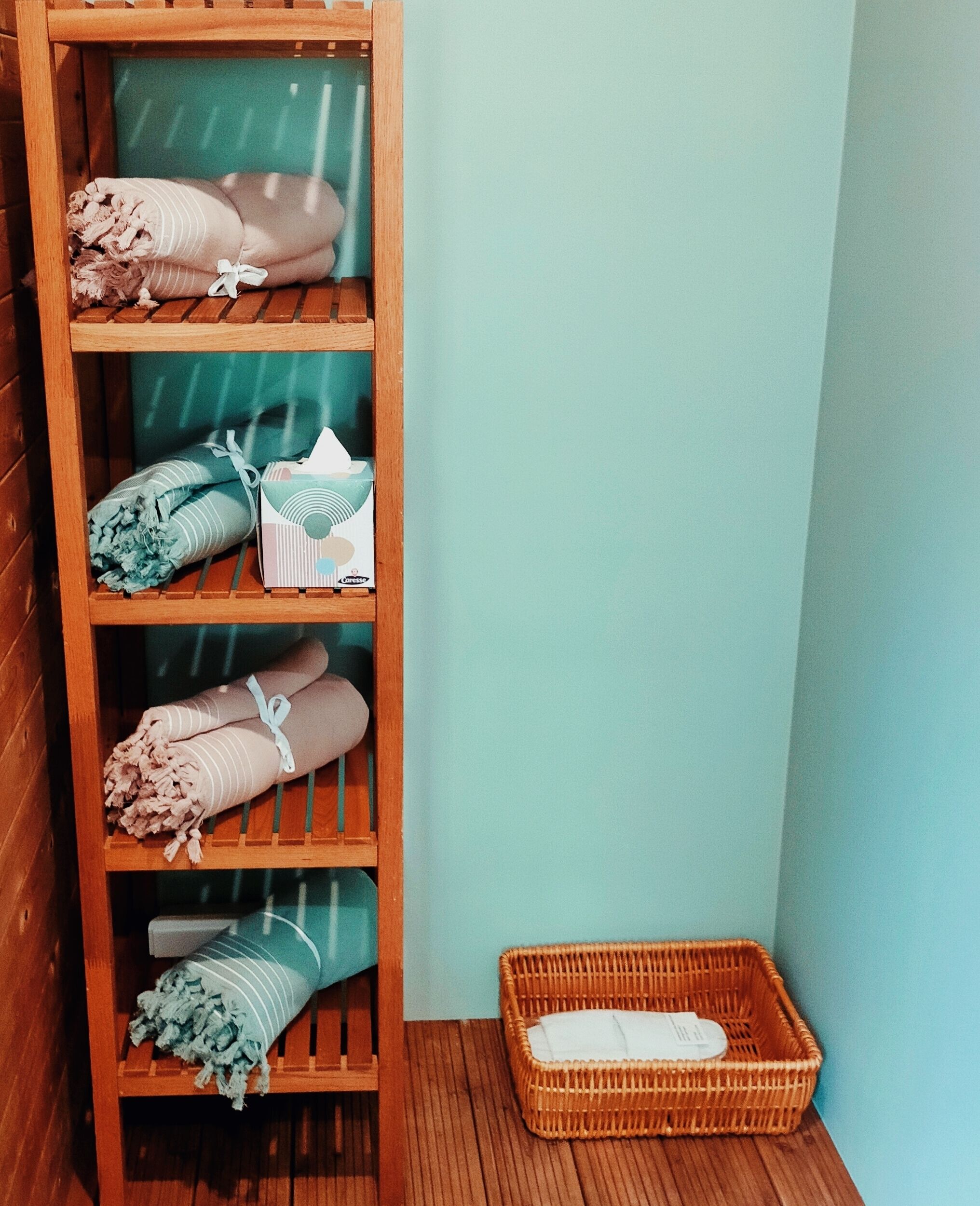Une étagère en bois avec des serviettes pliées et un panier, le tout posé sur un plancher en bois, devant un mur de couleur turquoise.