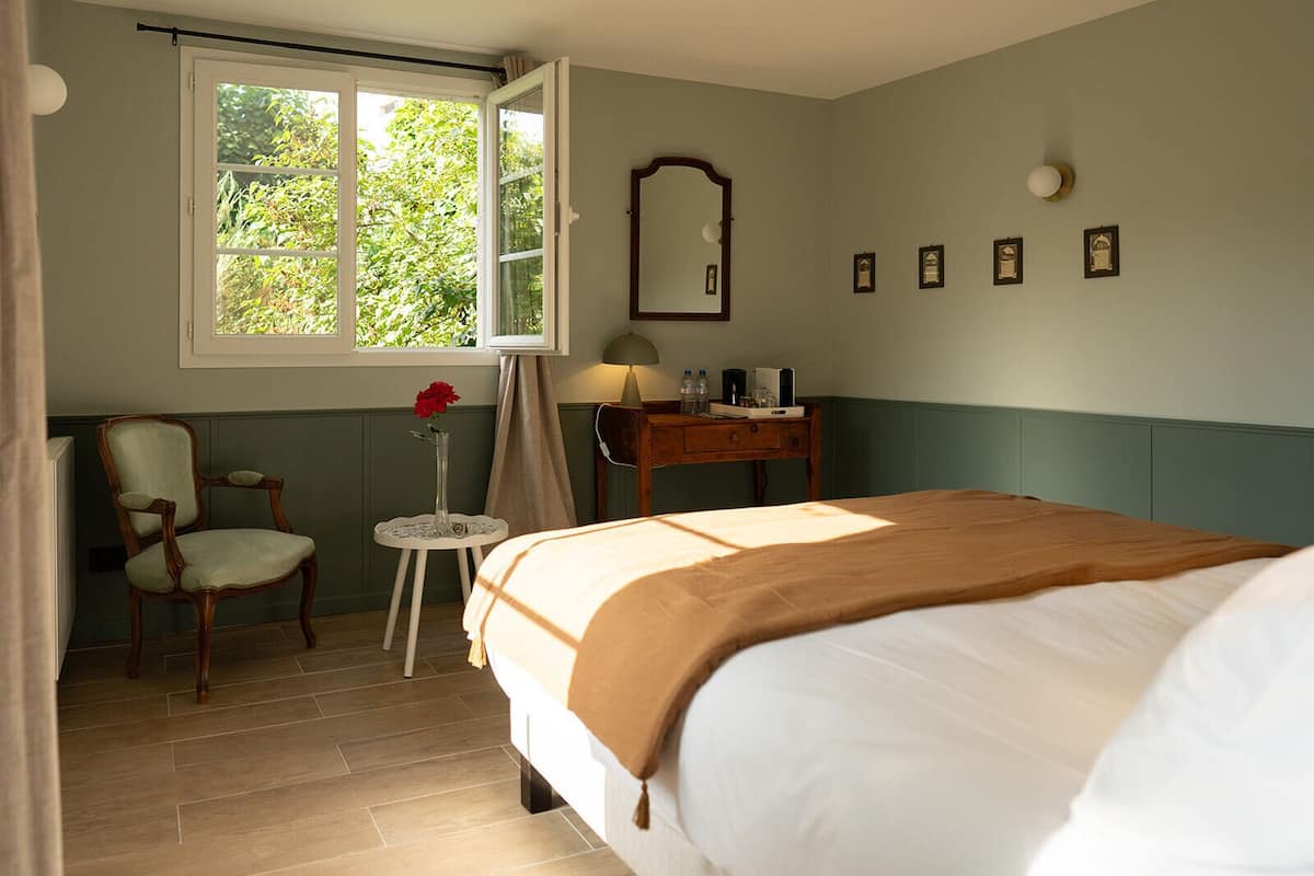 A bedroom with a bed, a chair, a small table with a vase of flowers, a lamp, and a window with a view of greenery.
