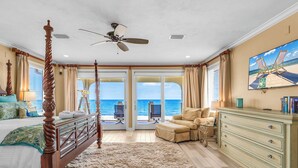 A spacious bedroom with a large bed, a ceiling fan, a TV, and a view of the ocean through the sliding doors.