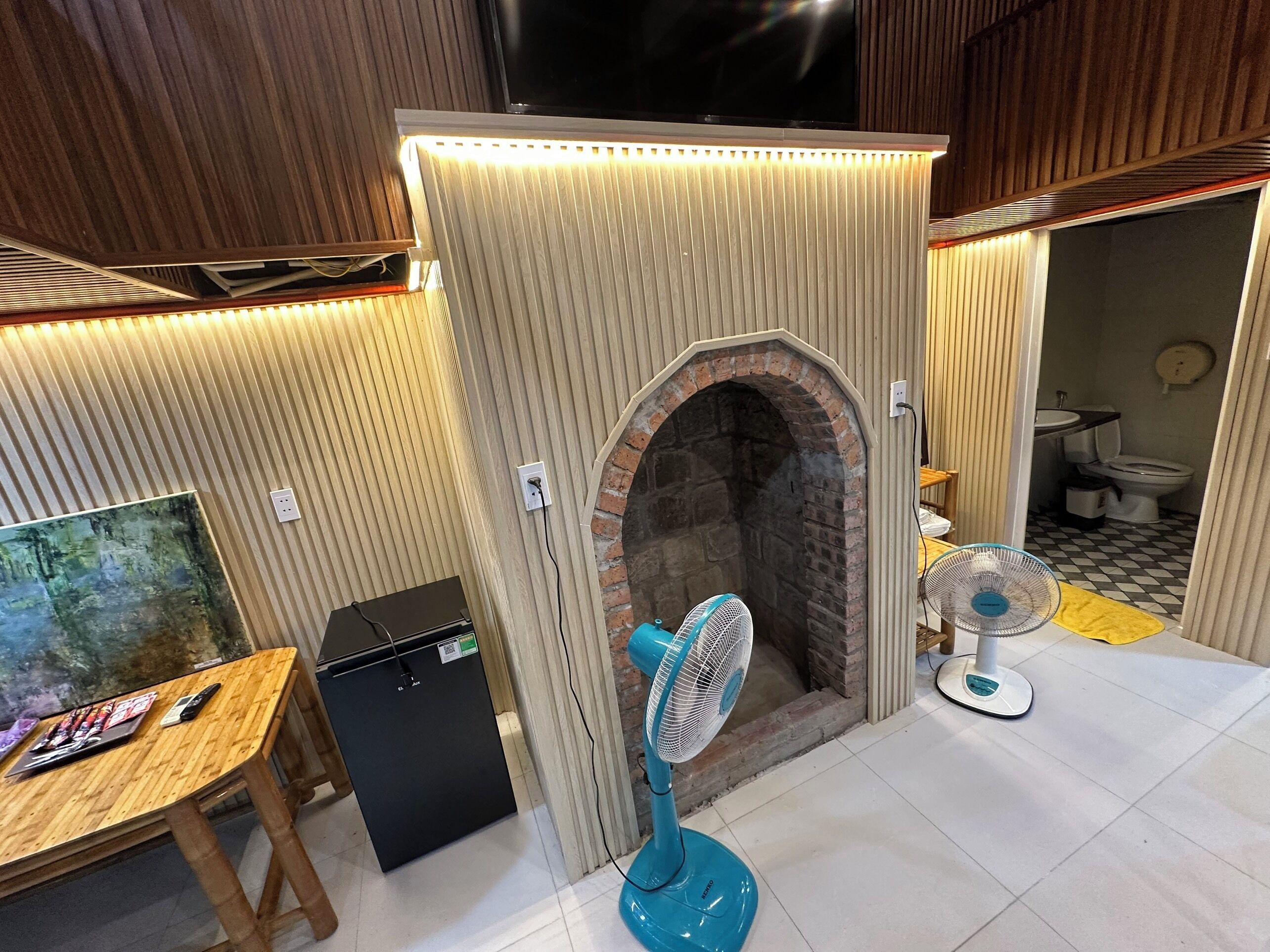 A room with a brick fireplace, a TV mounted above it, a small table with magazines, and a blue fan.