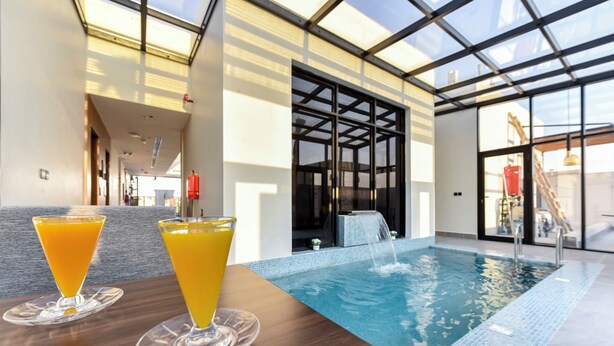 An indoor swimming pool with two glasses of orange juice on a table, large windows, and a view of the sky. - Marsal Park Hotel (Riyadh)
