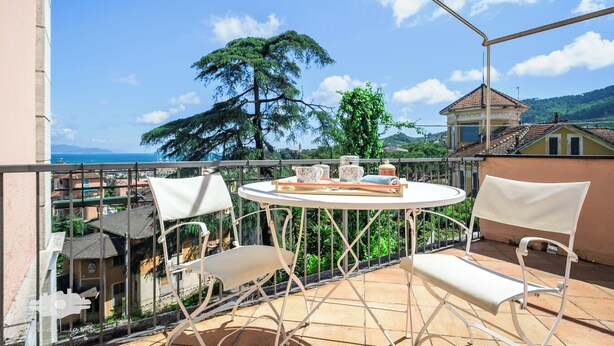Un balcón con una mesa redonda y dos sillas, con vista a un paisaje que incluye un árbol y edificios al lejos.