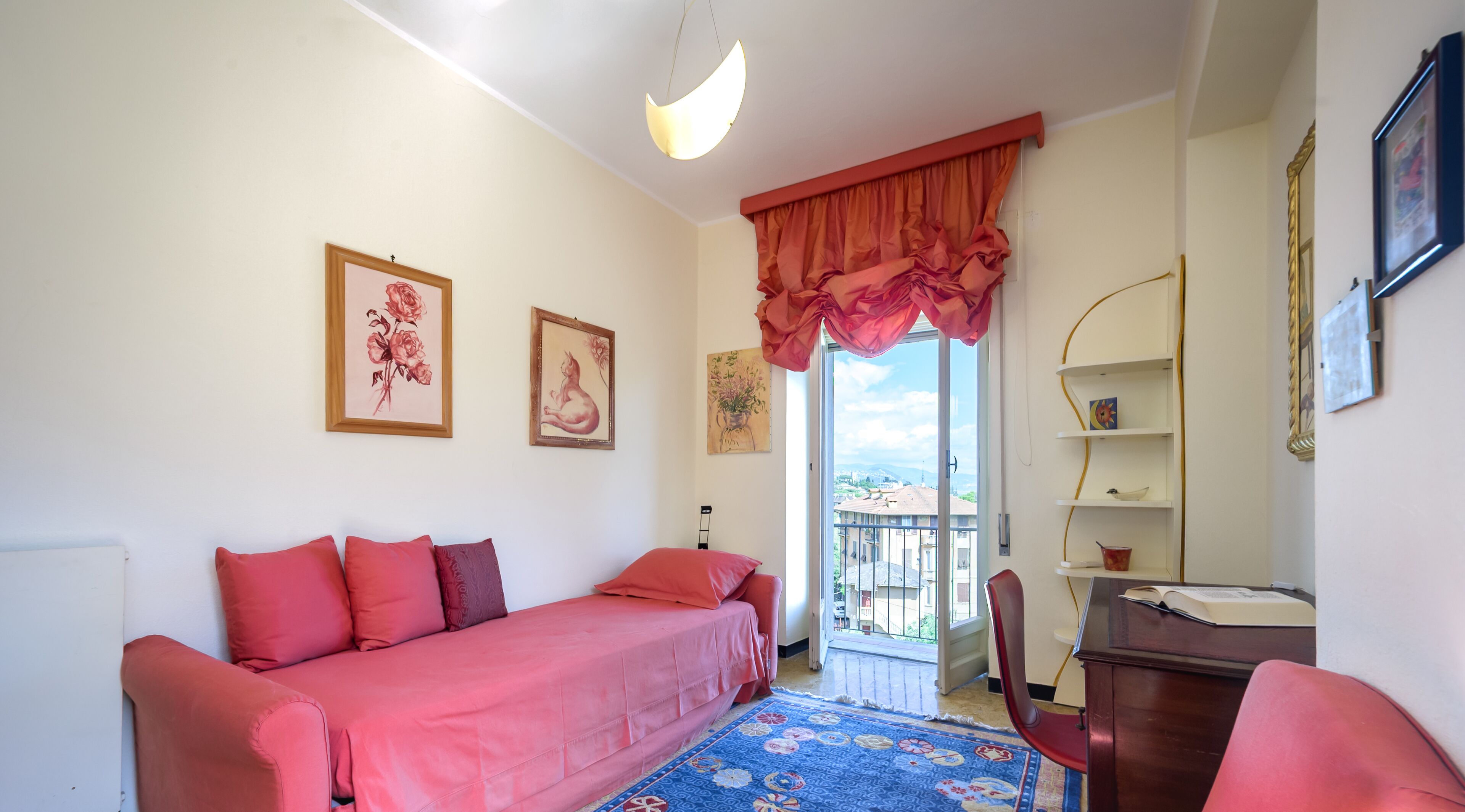 Une petite pièce agréable, dotée d’un lit, d’un bureau et d’une chaise. Les murs sont ornés de tableaux encadrés, il y a une fenêtre avec des rideaux et un balcon offrant une belle vue.
