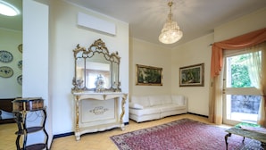 Un salón con un sofá blanco, una consola ornamentada y una araña. Hay dos cuadros en la pared y un ventanal con cortinas.