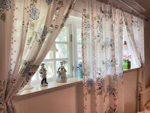 Una stanza con una finestra decorata da tende floreali, una piccola statuina sul davanzale e un vaso con fiori.