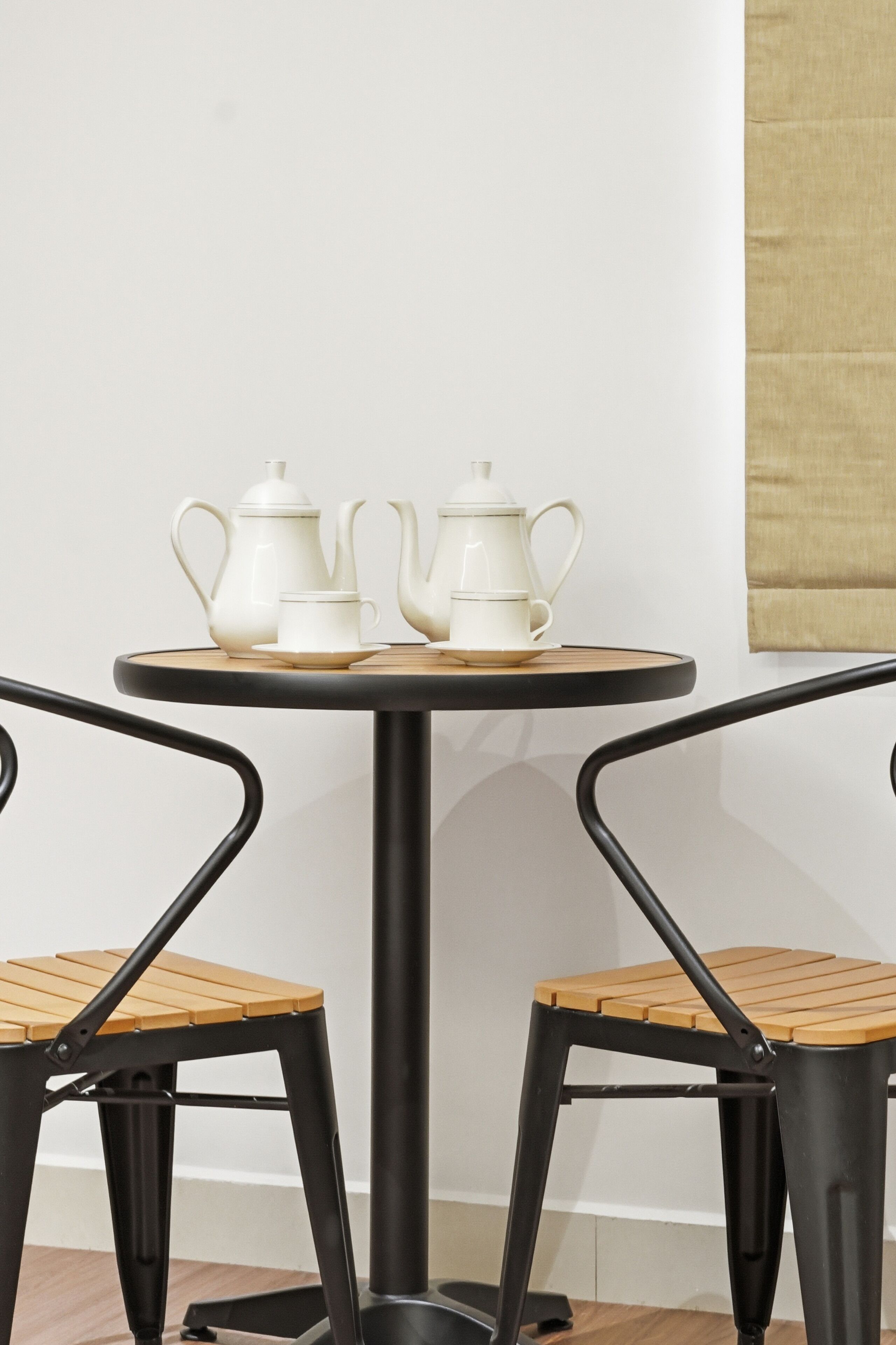 A round table with a white teapot, cup, and saucer, accompanied by a wooden tray. Two black chairs with wooden seats are positioned around the table.