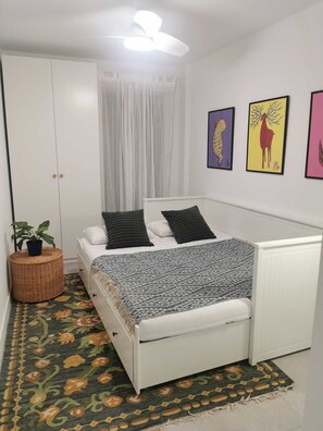A bedroom with a white bed, a small bedside table with a plant, a patterned rug, and three framed artworks on the wall.