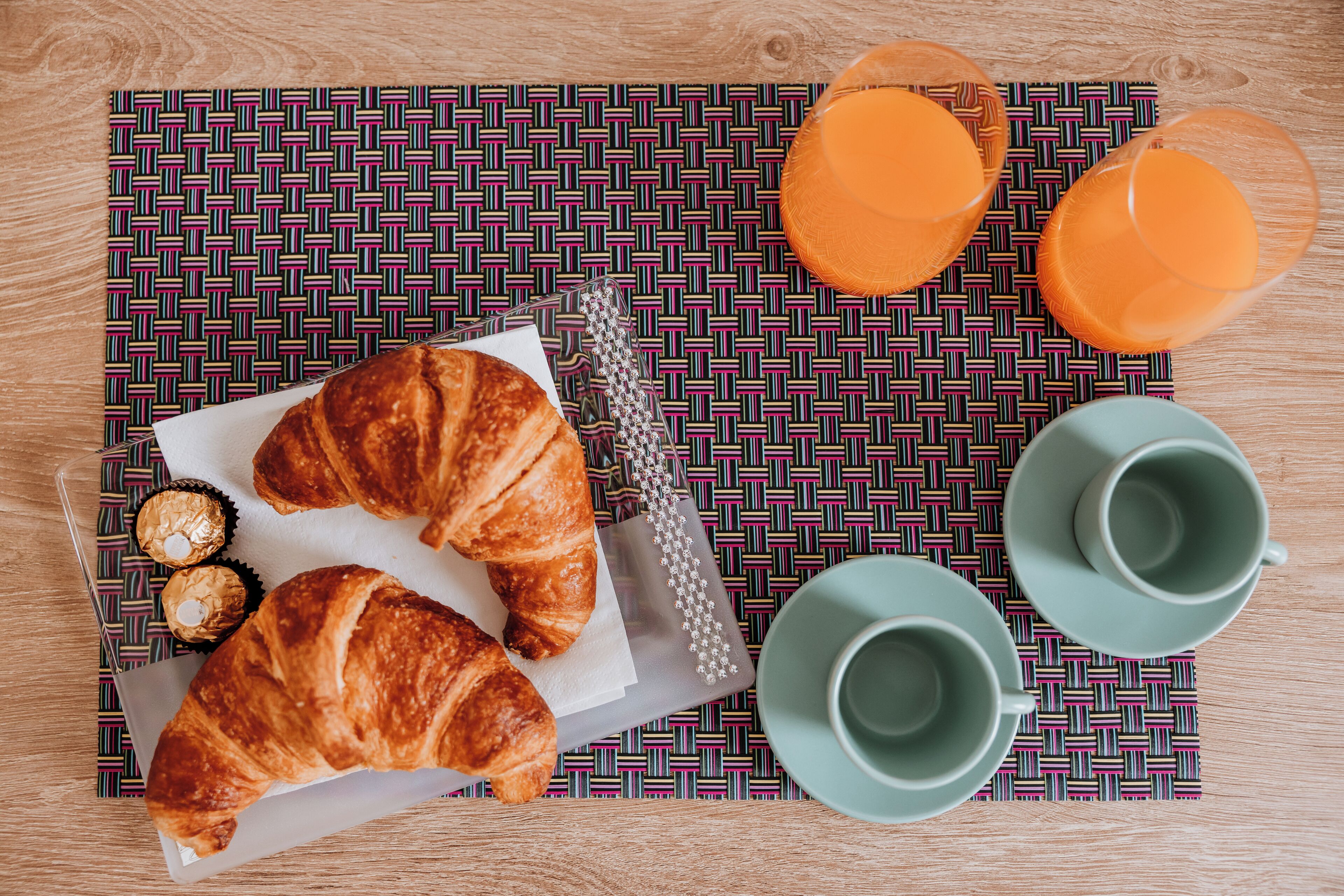 En frukostuppsättning med croissanter, två glas apelsinjuice och två gröna muggar placerade på en mönstrad servett.