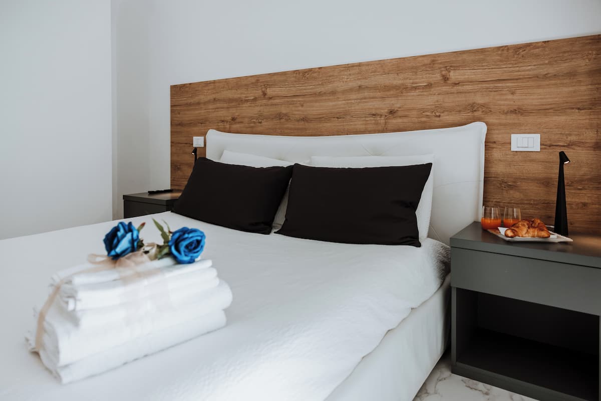 A neatly made bed with white linens and two black pillows, a wooden headboard, a bedside table with a lamp, and a tray with croissants and juice.