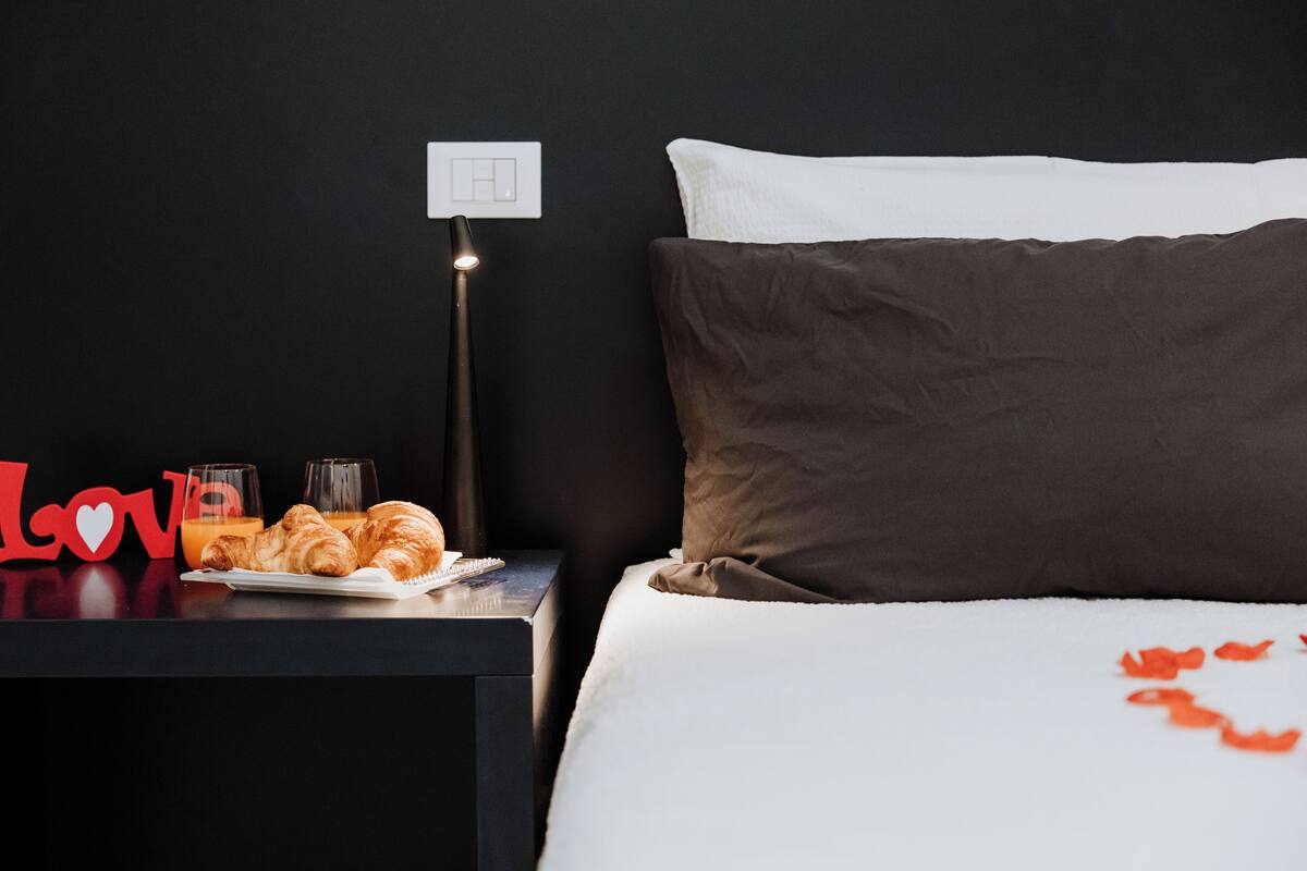 A neatly made bed with a white and brown pillow, a bedside table with a lamp, a plate of croissants, a glass of orange juice, and a red 'Love' sign.