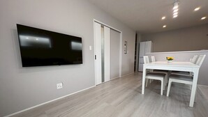 A modern, minimalist living room with a flat-screen TV mounted on the wall, a dining area with a white table and chairs, and a kitchen area with a refrigerator. - Yui-Takasaki (Takasaki)