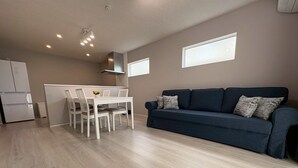 A modern living room with a dark blue sofa, a white dining table, and a kitchen area in the background. - Yui-Takasaki (Takasaki)