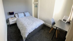 Una camera da letto con un letto, un comodino, una sedia e una piccola scrivania con un quadro incorniciato.