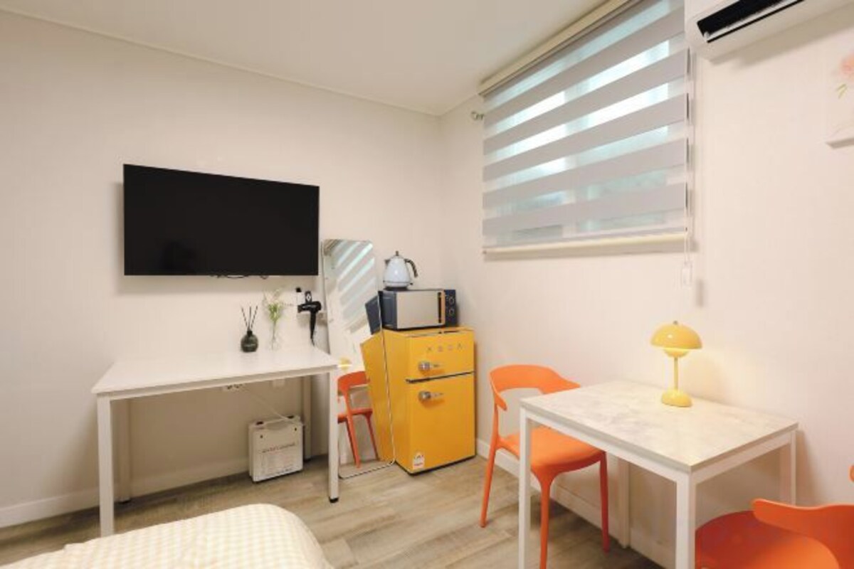 A room with a bed, a desk, a TV, a small cabinet, and a window with blinds.