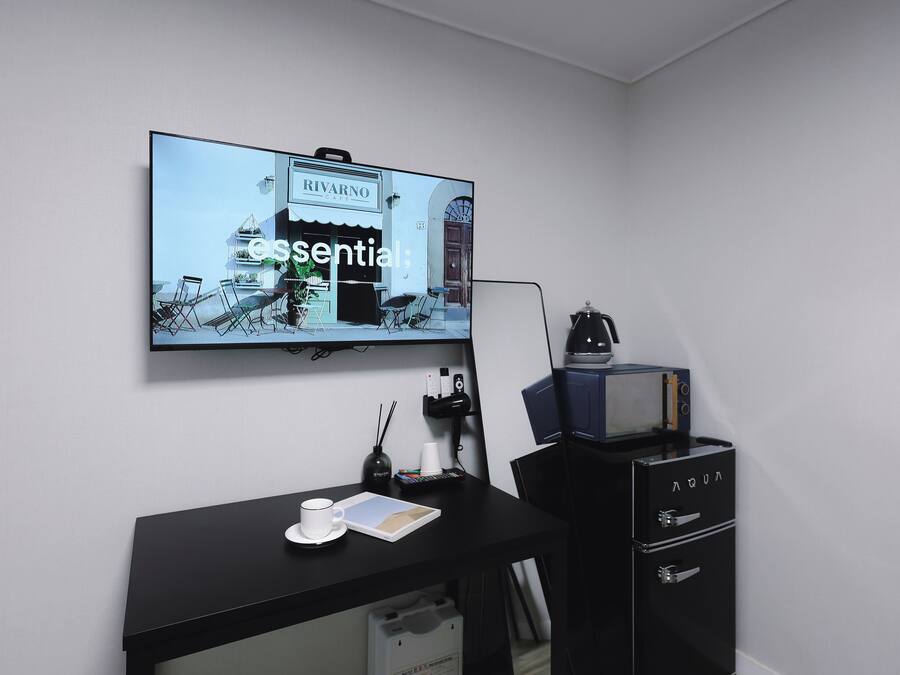 Una habitación con una televisión de pantalla plana montada en la pared mostrando una escena de café, un escritorio negro con una taza y un bloc de notas, y un archivador negro con cajones.
