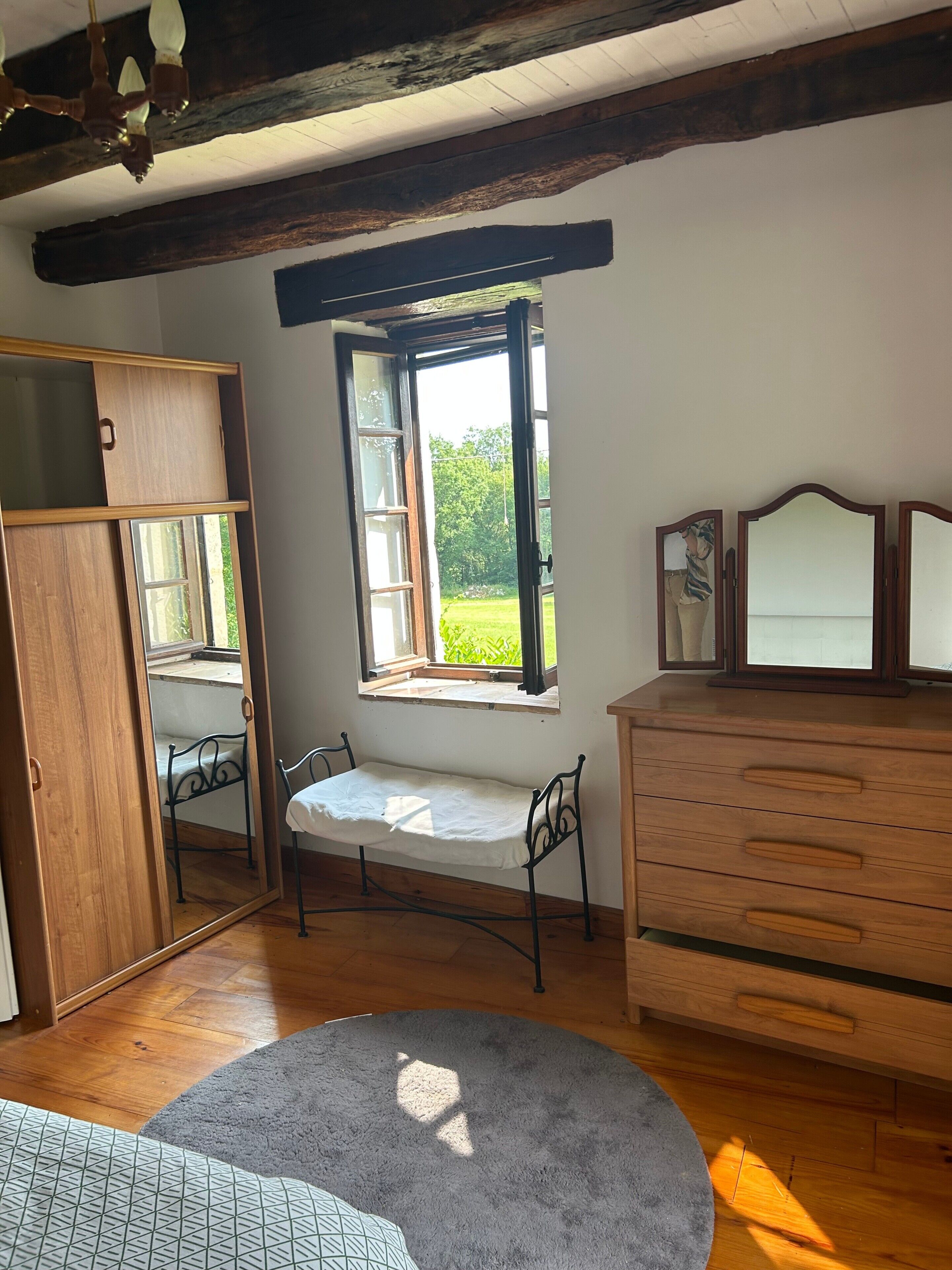 A room with a wooden wardrobe, a bench, a dresser, and a window with a view of greenery.