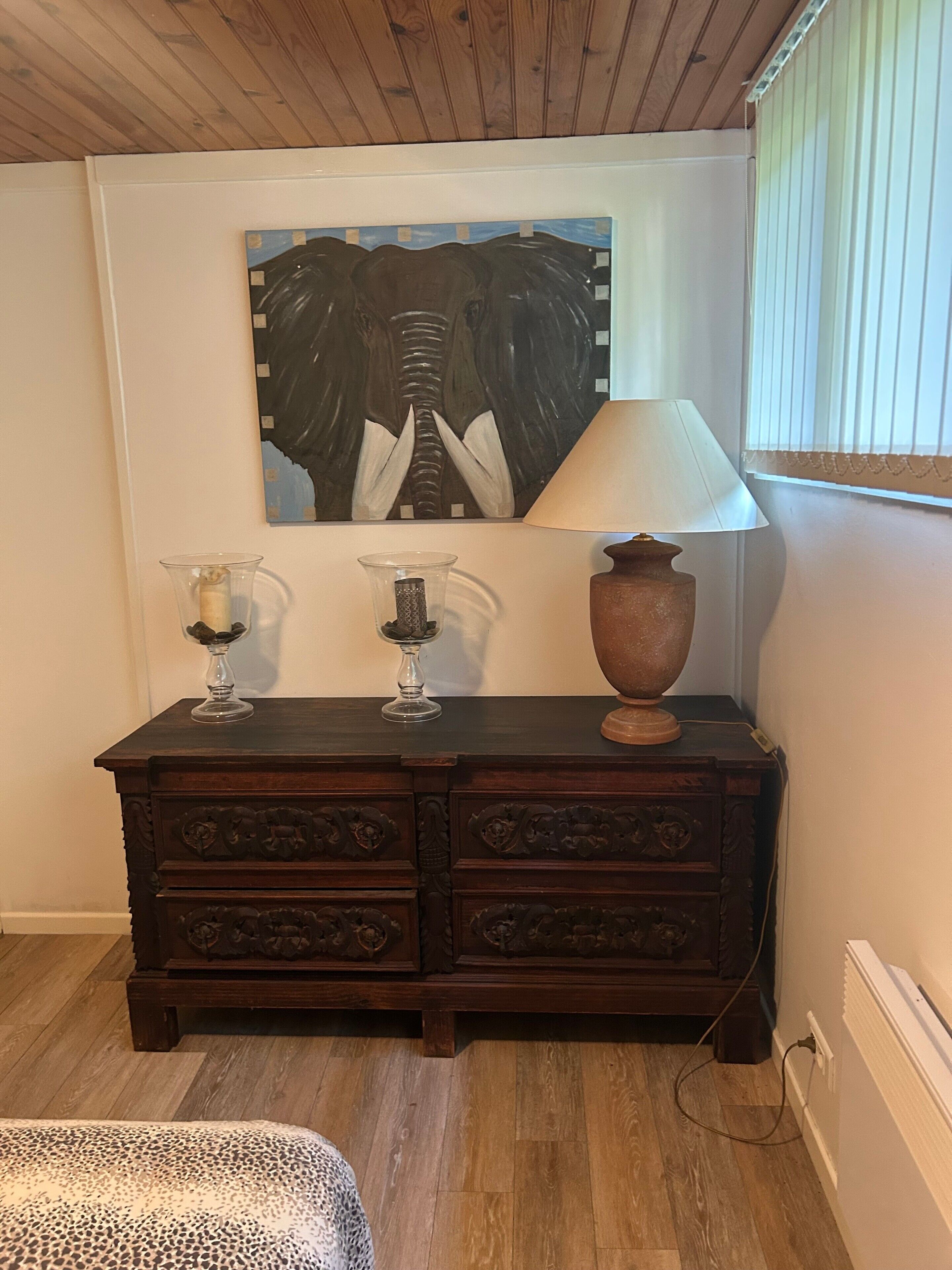 A wooden dresser with two glass vases and a lamp, a painting on the wall, and a radiator.