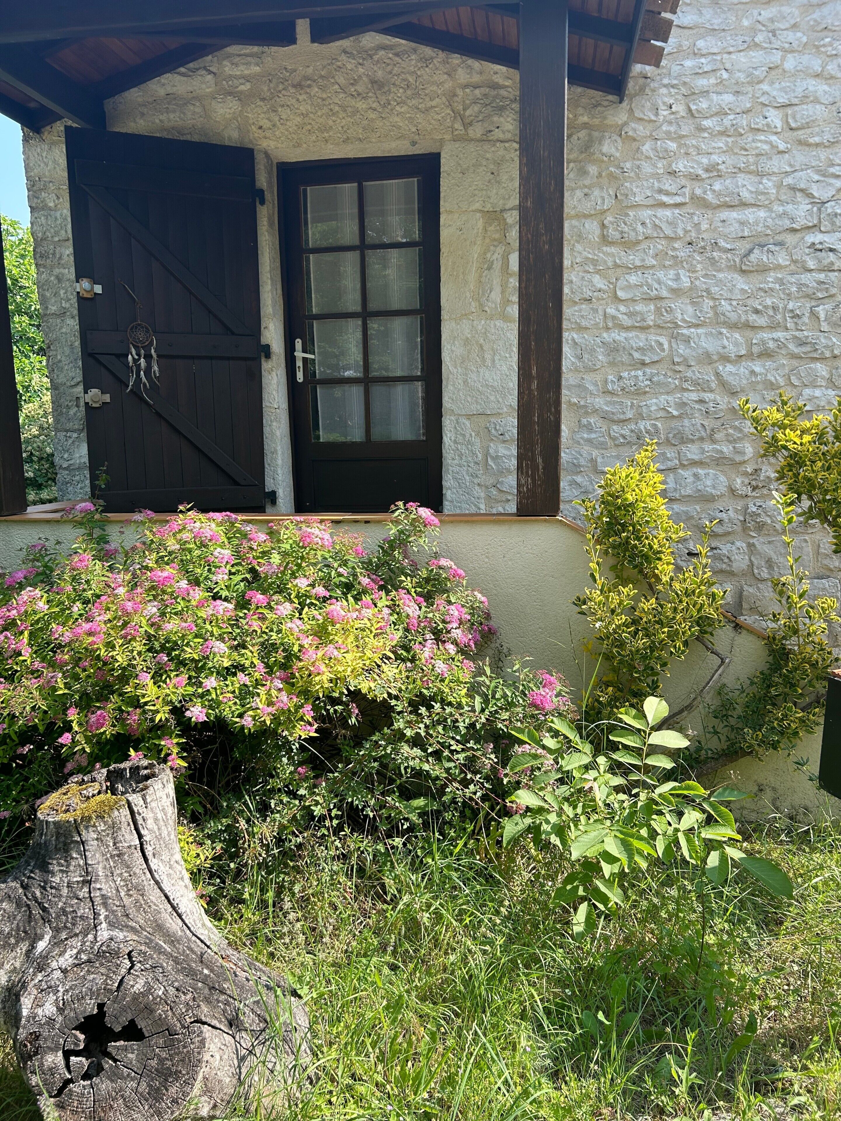 A stone building with a dark wooden door, a window, and a small porch area with pink flowers and greenery.