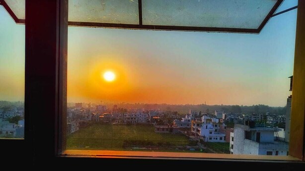 Une vue par une fenêtre au coucher du soleil, avec un paysage urbain comprenant des bâtiments et un ciel dégagé.